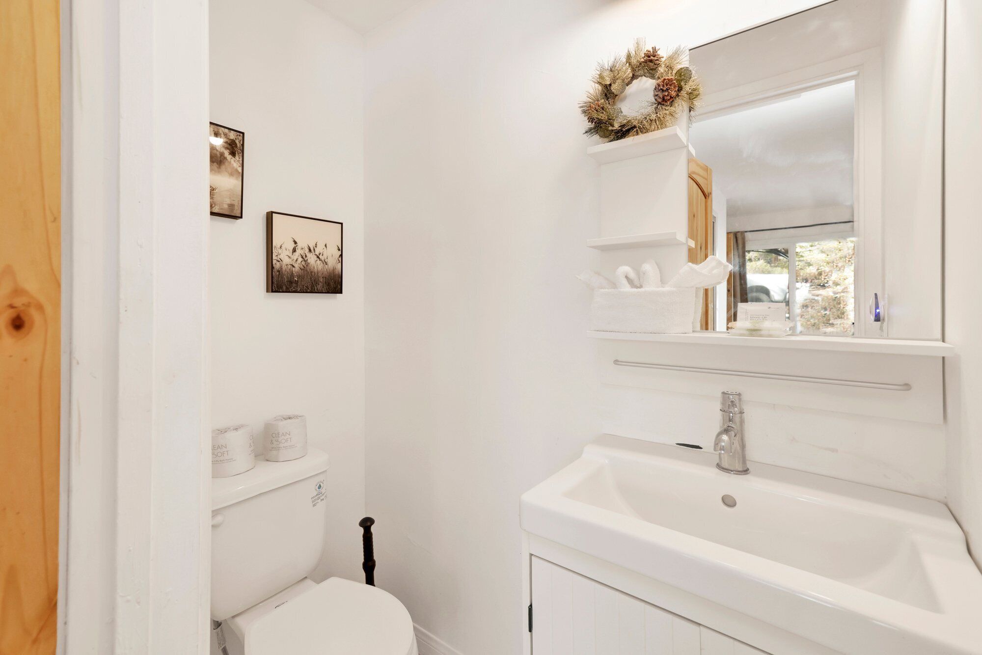 A bathroom with a toilet, a sink, and a mirror. There are two framed pictures on the wall, a wreath on the mirror, and a tissue box on the sink.