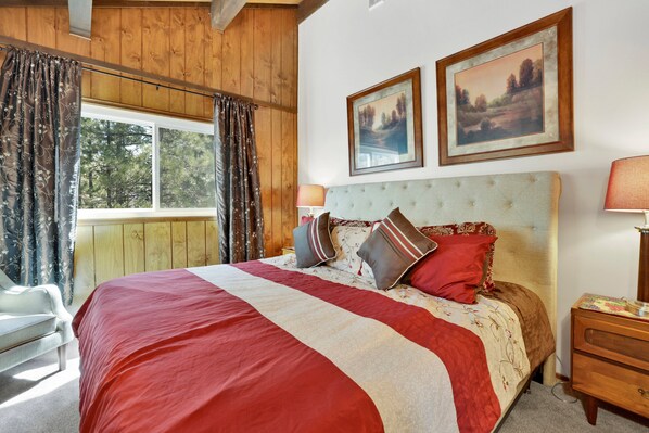 Un dormitorio con una cama grande, vigas de madera en el techo, una ventana con cortinas y dos cuadros en la pared.