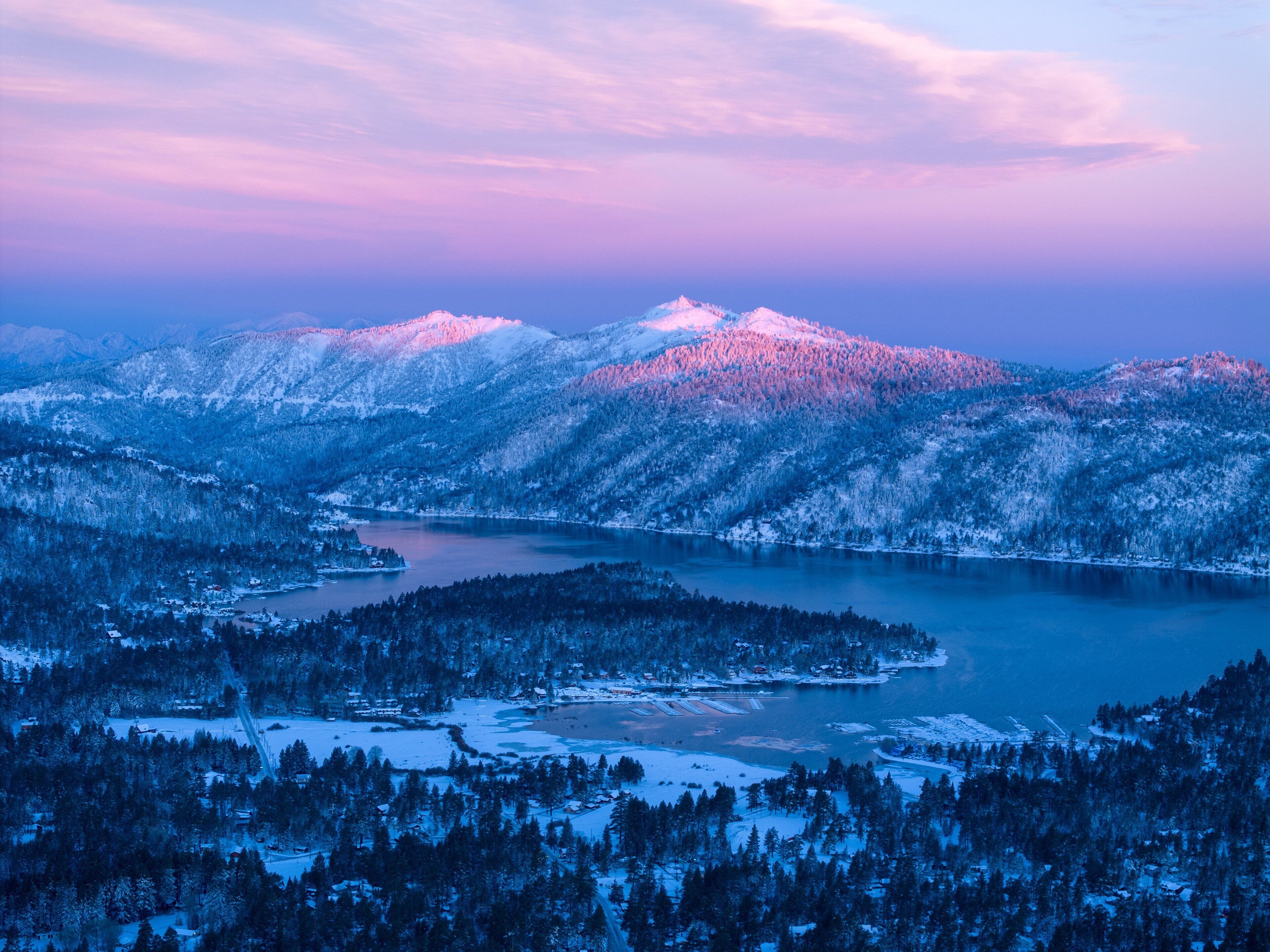 A panoramic view of a snow-covered landscape with a lake, mountains, and a small settlement.