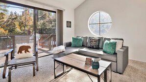 A modern living room with a grey sofa, a coffee table, and a chair with a bear pillow. There's a large window with a circular stained glass design, and a dartboard on the wall. - Restmoore #2425 (Big Bear Lake)