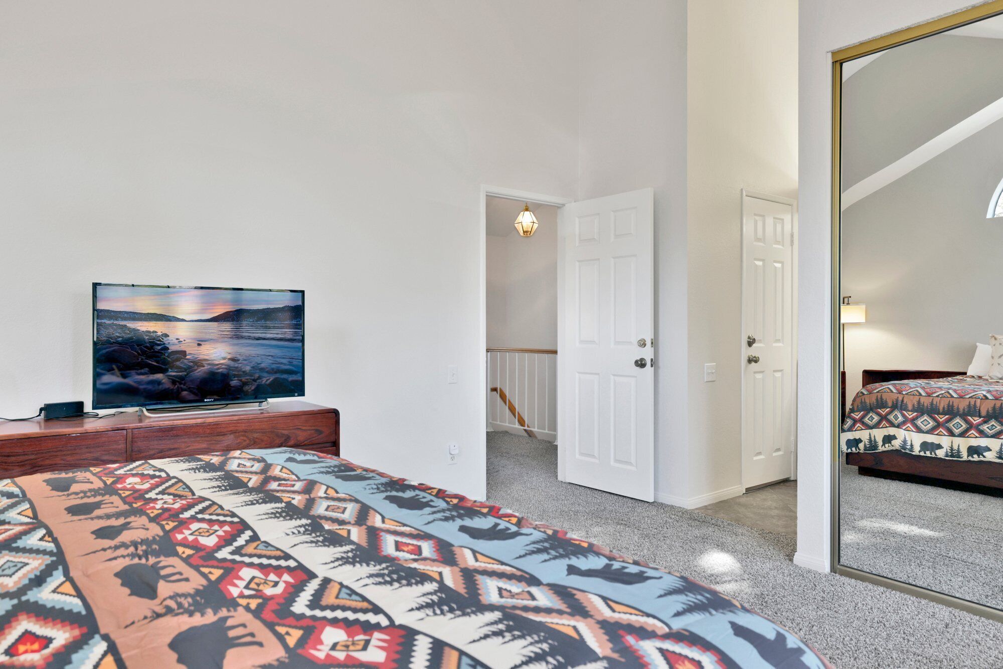 Un dormitorio con una cama, un televisor sobre un aparador de madera, un tapete estampado y un espejo grande que refleja la habitación.