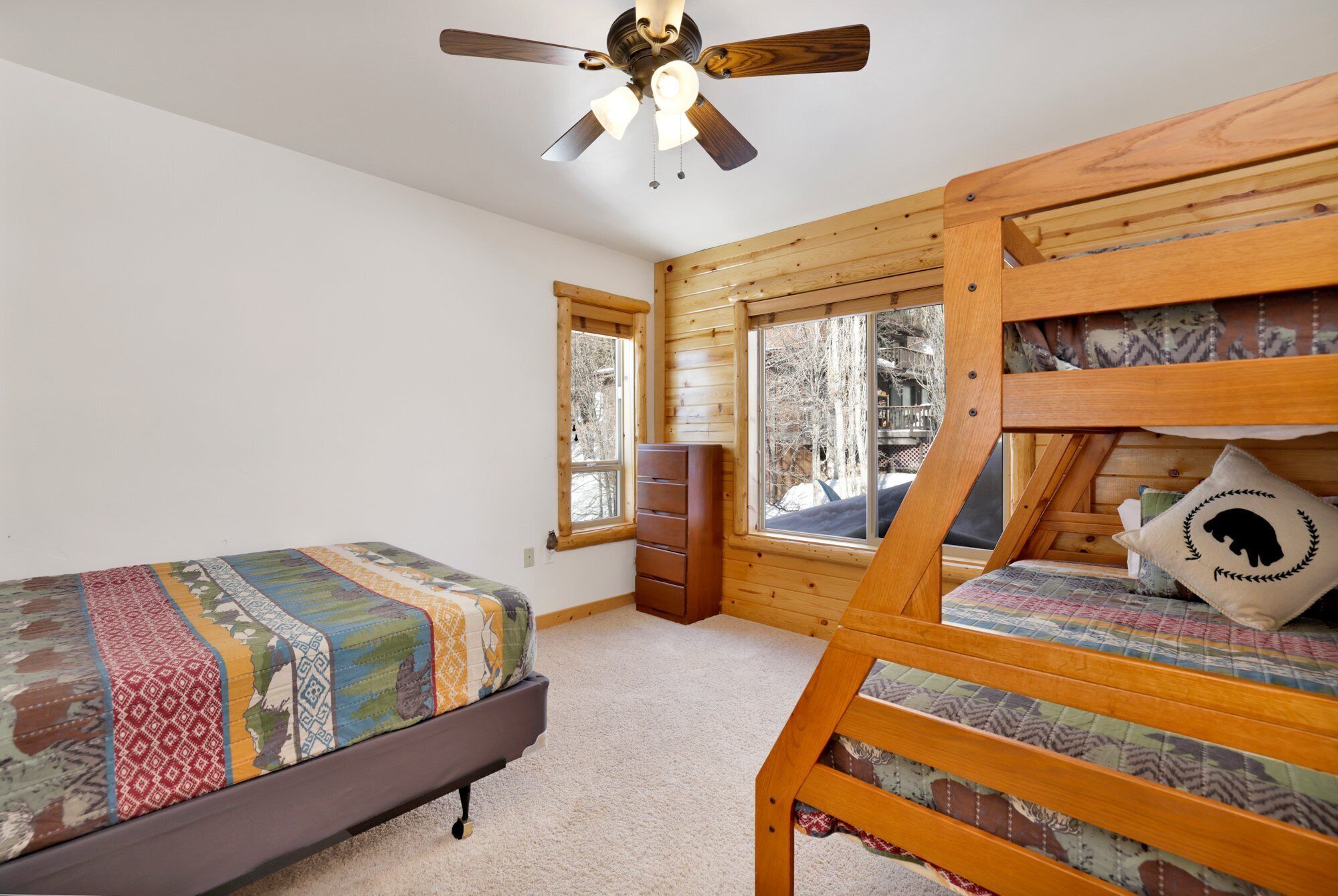 A cozy bedroom with a bunk bed, a single bed, a ceiling fan, and a window with a view of trees.
