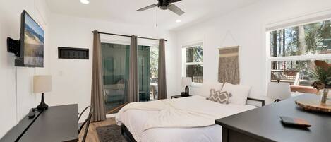 A modern bedroom with a bed, a desk, a ceiling fan, and a large window.
