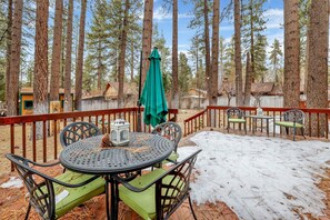 A deck with a round table and chairs, surrounded by tall trees and a wooden fence. - Flitterbick Lodge #2246 (Fawnskin)