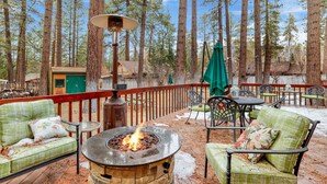 An outdoor patio with a stone fire pit, green cushioned chairs, and a wooden deck. - Flitterbick Lodge #2246 (Fawnskin)