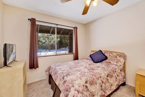 Une chambre à coucher avec un lit, une fenêtre avec des rideaux, un ventilateur de plafond et un téléviseur posé sur un meuble.