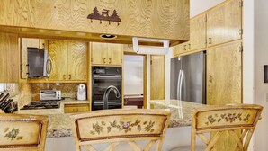 Une cuisine dotée de meubles en bois, d’un four encastré, d’un réfrigérateur et d’une machine à café. On y trouve deux chaises recouvertes d’un tissu à motifs floraux.