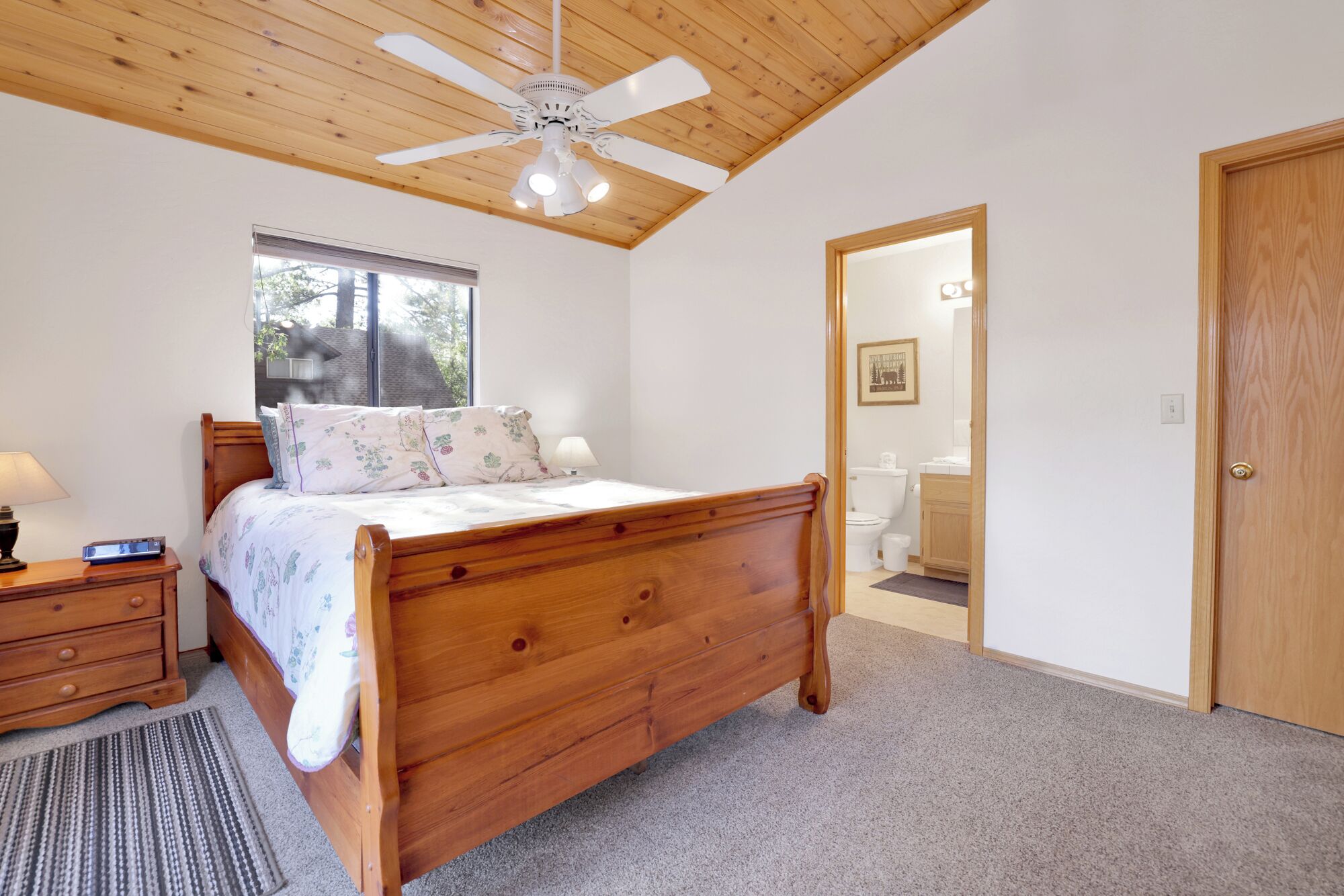 Une chambre à coucher avec un lit en bois, un ventilateur de plafond, une fenêtre et une porte donnant sur une salle de bain.
