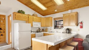 Une cuisine avec des meubles en bois, un réfrigérateur blanc, un four à micro-ondes et une cuisinière. Il y a des tabourets près de l’îlot central et un extincteur est fixé au mur.