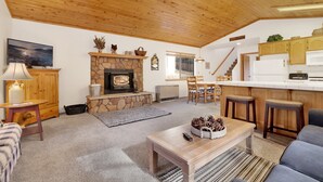 Un salon chaleureux avec une cheminée en pierre, un plafond en bois et un coin cuisine doté de meubles en bois et d’un réfrigérateur blanc.