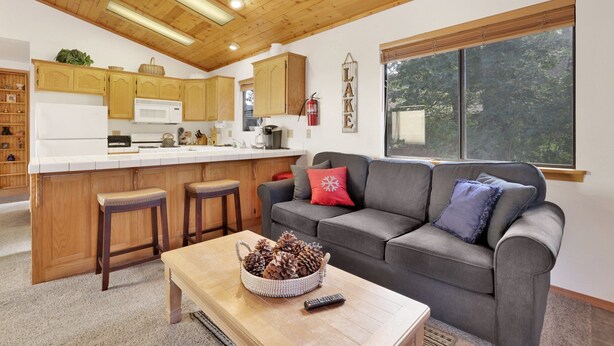 Une cuisine moderne avec des meubles en bois et un îlot central, un salon confortable avec un canapé gris et une table basse, et une grande fenêtre donnant sur la verdure.