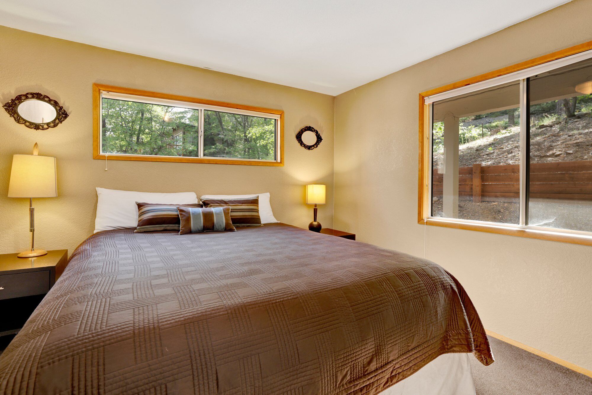 A bedroom with a large bed, two bedside lamps, a window with a view of trees, and a mirror on the wall.