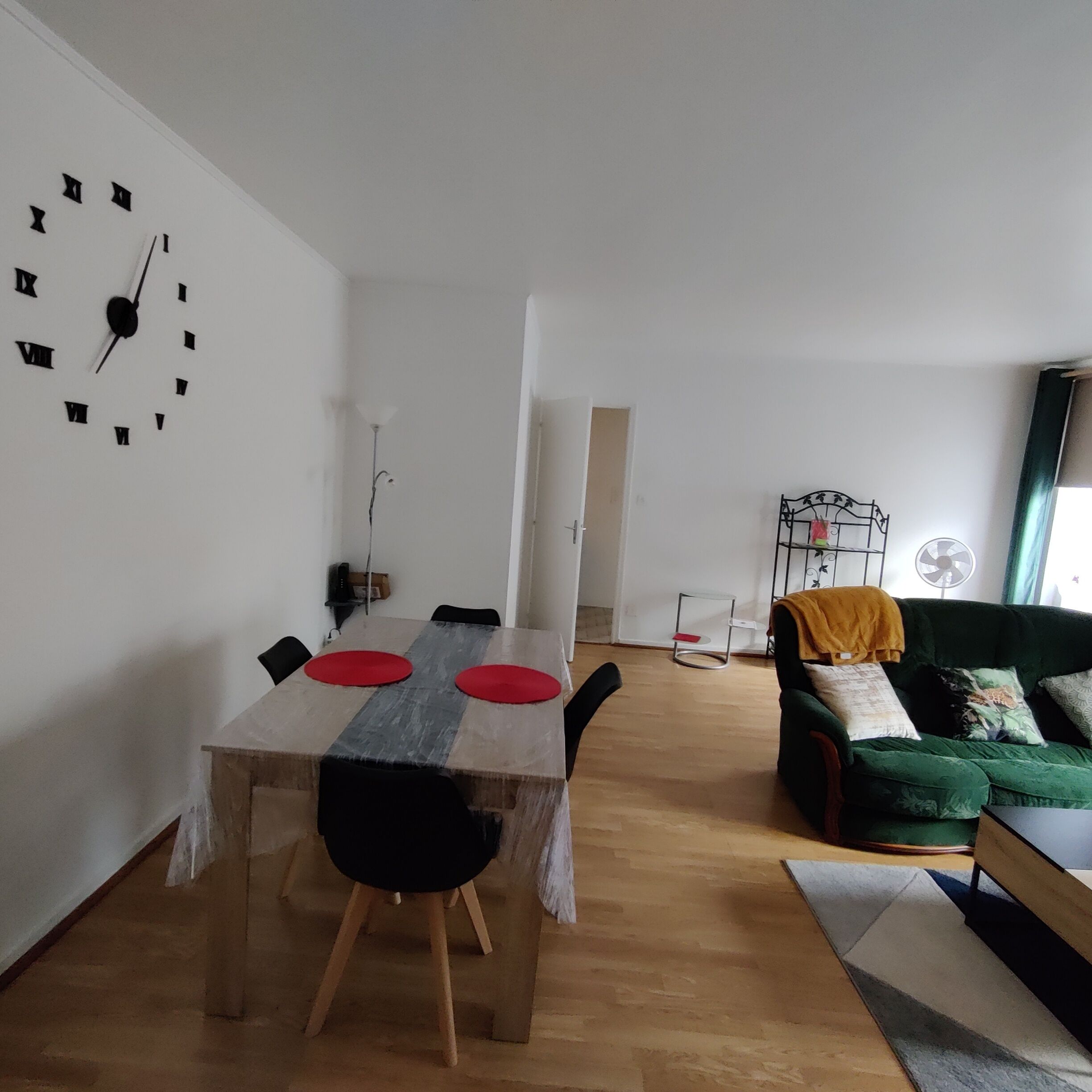 Un comedor con una mesa de madera, sillas y un reloj de pared. Se ve un sofá verde y un ventilador al fondo.