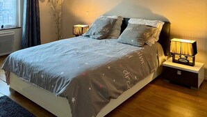 Een slaapkamer met een bed, nachtlampjes, een raam met gordijnen, een wanddecoratie en een klein tafeltje met een lamp.