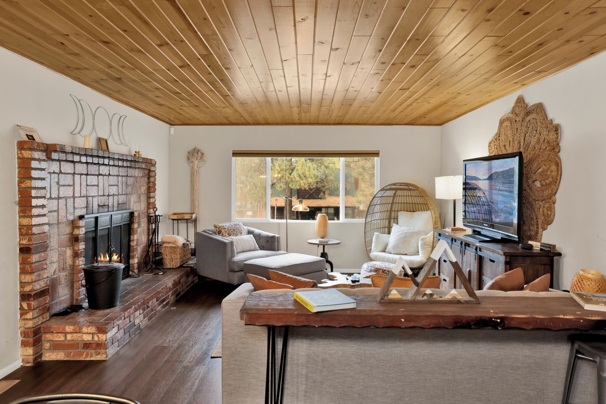 A cozy living room with a brick fireplace, a large window, a television, and a wooden ceiling.