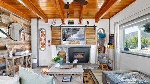 A cozy living room with a fireplace, wooden ceiling, and a large window overlooking a scenic view.
