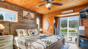 A wooden cabin bedroom with a bed, nightstands, a ceiling fan, a large window, and a view of the outdoors.