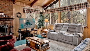 Interior acollidor d'una cabana amb una llar de foc, un televisor que mostra un logotip, un sofà gris i una finestra gran amb cortines escoceses.