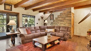 A cozy living room with a large brown leather sofa, a wooden coffee table, and a red shag rug. A stone wall and wooden beams are visible in the background. - HOW Sweet IT IS #1531 (Big Bear Lake)