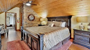 A bedroom with a stone fireplace, a bed with striped bedding, a wooden ceiling fan, and a nightstand with a lamp. - HOW Sweet IT IS #1531 (Big Bear Lake)