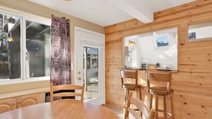 Una habitación con paneles de madera, una mesa redonda para comer, dos sillas de madera, una ventana con cortinas, y una pequeña zona de cocina con un espejo y un cuadro en la pared.