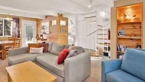 Una sala de estar acogedora con un sofá gris, un sillón azul y una mesa de centro de madera. Se ve una escalera con barandilla de madera que conduce a un nivel superior.