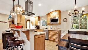 A modern kitchen with wooden cabinets, a stainless steel range hood, a microwave, and a dining area with a wooden table and bar stools. - A Hidden Asset #2379 (Big Bear Lake)
