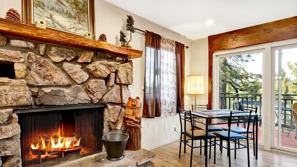 A stone fireplace with a fire, a dining table with chairs, and a balcony with a view of trees. - A Hidden Asset #2379 (Big Bear Lake)