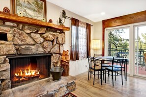 A stone fireplace with a fire, a dining table with chairs, and a balcony with a view of trees. - A Hidden Asset #2379 (Big Bear Lake)