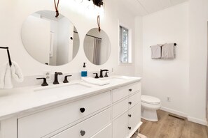 A modern bathroom with a white vanity, two round mirrors, a white toilet, and a window providing natural light.