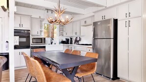 A modern kitchen with white cabinetry, a stainless steel refrigerator, a dining table with brown chairs, and a unique antler chandelier.
