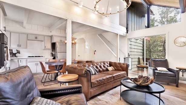 A spacious living area with a leather sofa, a coffee table, and a dining table with chairs. There is a kitchen in the background with white cabinets and a stainless steel refrigerator.