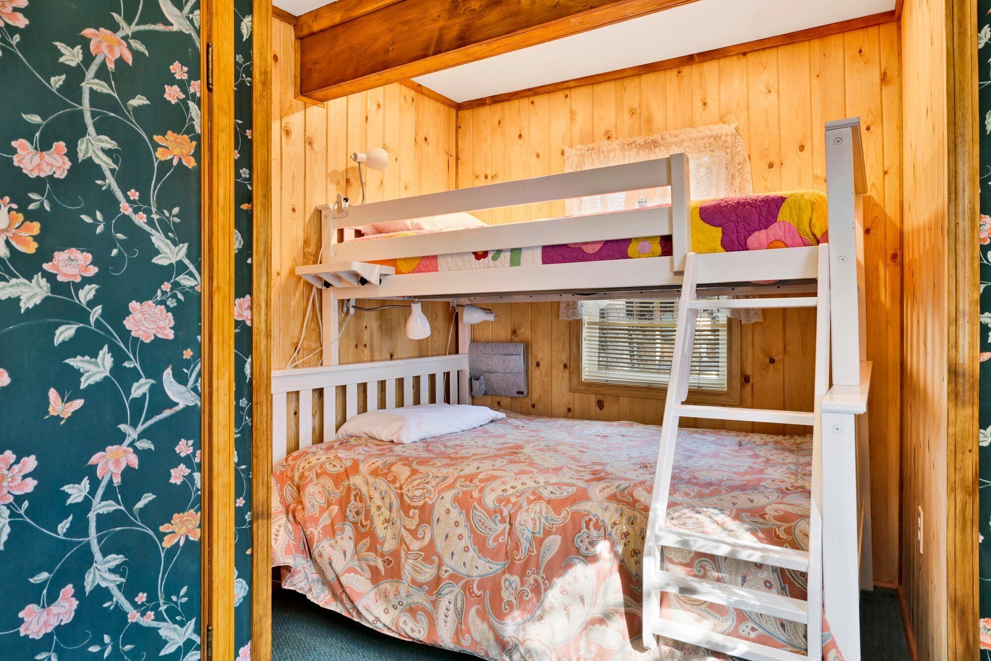 Une chambre avec un lit superposé, un plafond et des murs en bois, une échelle, une fenêtre avec des stores et un papier peint à motifs floraux sur un des murs.