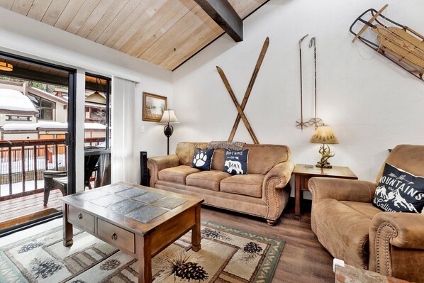 A cozy living room with a wooden ceiling, a brown leather sofa, a wooden coffee table, and a patterned rug.