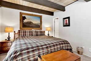A bedroom with a bed, two bedside tables, a nightstand, a wooden bench, a painting, and a framed picture on the wall.