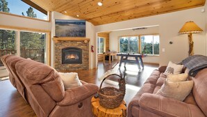 A cozy living room with a fireplace, wooden ceiling, and large windows offering a view of the outdoors. - Silver TIP Chalet #2141 (Big Bear Lake)