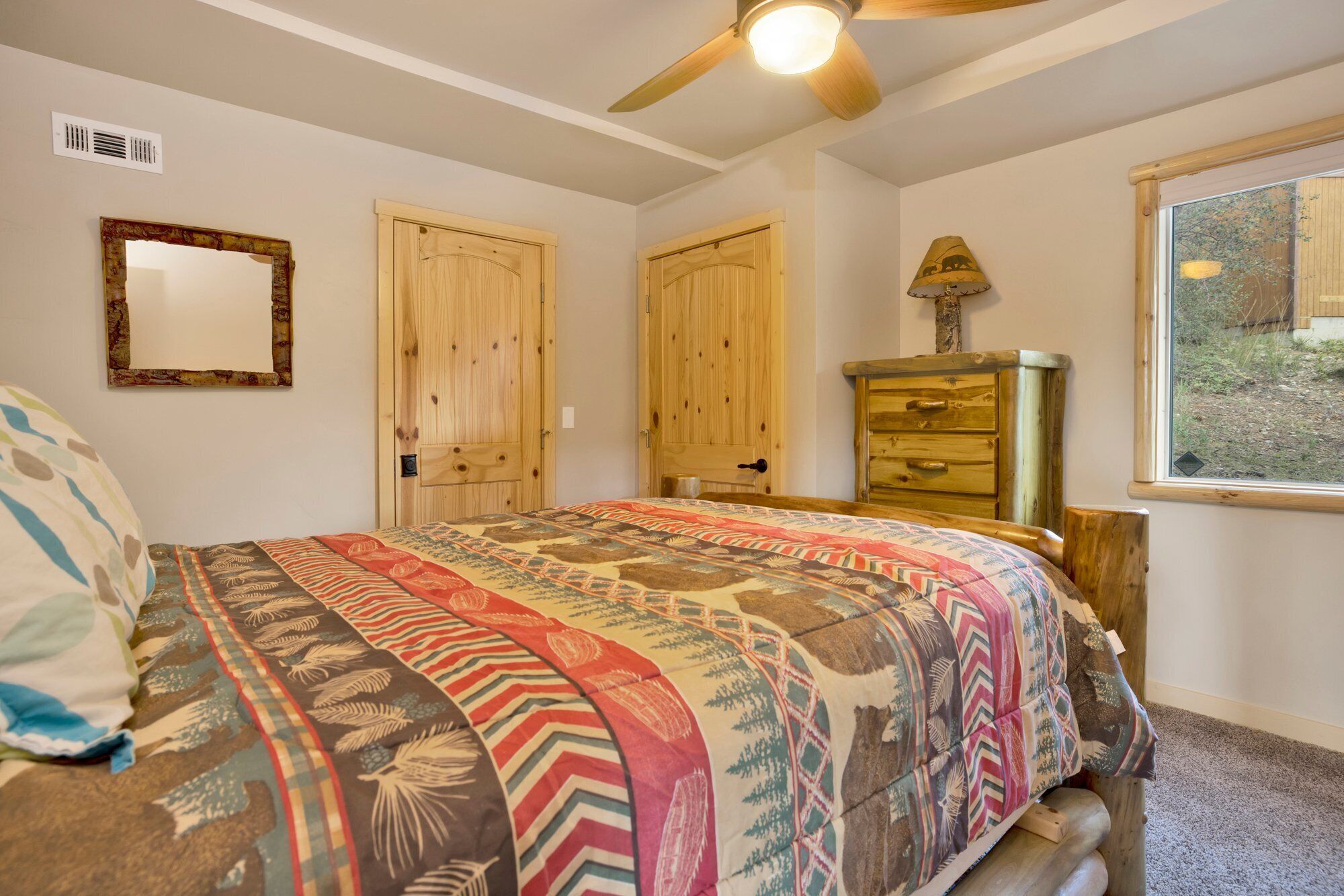 A bedroom with a bed, wooden doors, a dresser, a ceiling fan, and a window with a view of greenery.
