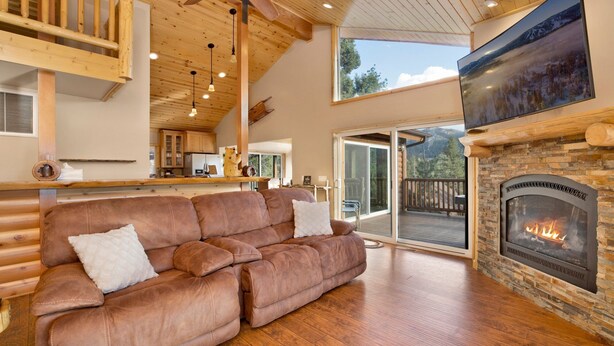A cozy living room with a fireplace, wooden flooring, and a large window overlooking a deck. - Silver TIP Chalet #2141 (Big Bear Lake)