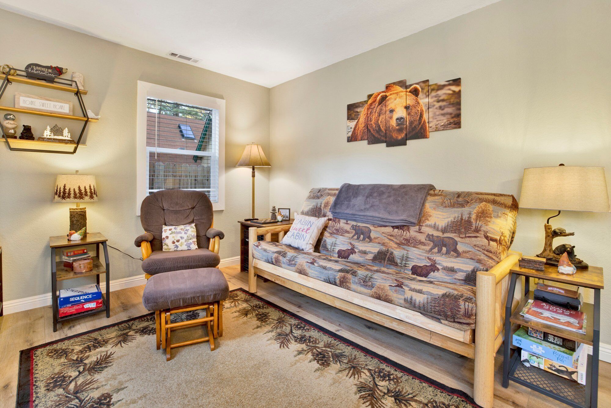 A cozy living room with a sofa, armchair, and a rug. There are bookshelves, a lamp, and a window with blinds.