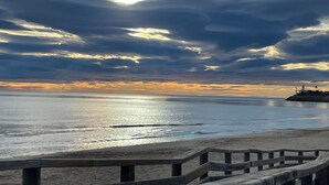 Ein Strand mit einer Holzpromenade, die zum Wasser führt, einem Leuchtturm in der Ferne und einem Sonnenuntergang mit Wolken.