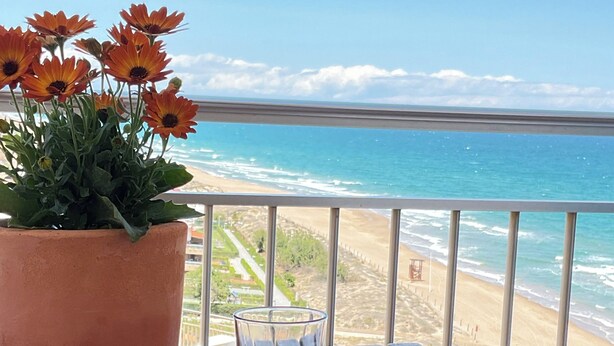 Ein Balkon mit Esstisch, einem Blumentopf mit Blumen und Blick auf Strand und Meer.