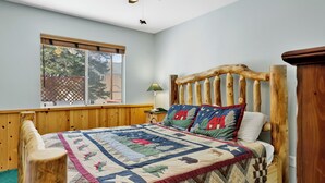 A bedroom with a wooden headboard, a quilt with a house pattern, a nightstand with a lamp, and a window with blinds. - Alameda Landing #2345 (Big Bear City)