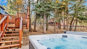 A hot tub with a wooden deck and a wooden staircase leading to a deck area surrounded by trees. - Alameda Landing #2345 (Big Bear City)