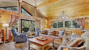 L’intérieur d’un chalet accueillant, avec un plafond en bois, de grandes fenêtres, un canapé, un fauteuil, une table et une lampe.