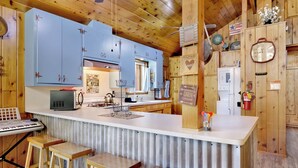 Une cuisine de style rustique, avec des meubles bleus, un réfrigérateur blanc, une bouilloire et une machine à café. On y trouve trois tabourets en bois et un évier en métal.