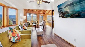 A spacious living area with a large window, wooden ceiling beams, a ceiling fan, and a painting of a lake. - THE Octagon #2394 (Big Bear City)