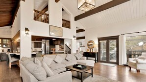 A spacious living area with a large sectional sofa, a coffee table, and a dining area with a table and chairs. There is a kitchen visible in the background, and a staircase leading to an upper level. - Soaring Pines #2193 (Big Bear Lake)