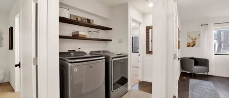 Een moderne keuken met twee Maytag wasmachines, een houten plank erboven en een zithoek met een grijze bank en een schilderij van een hert.
