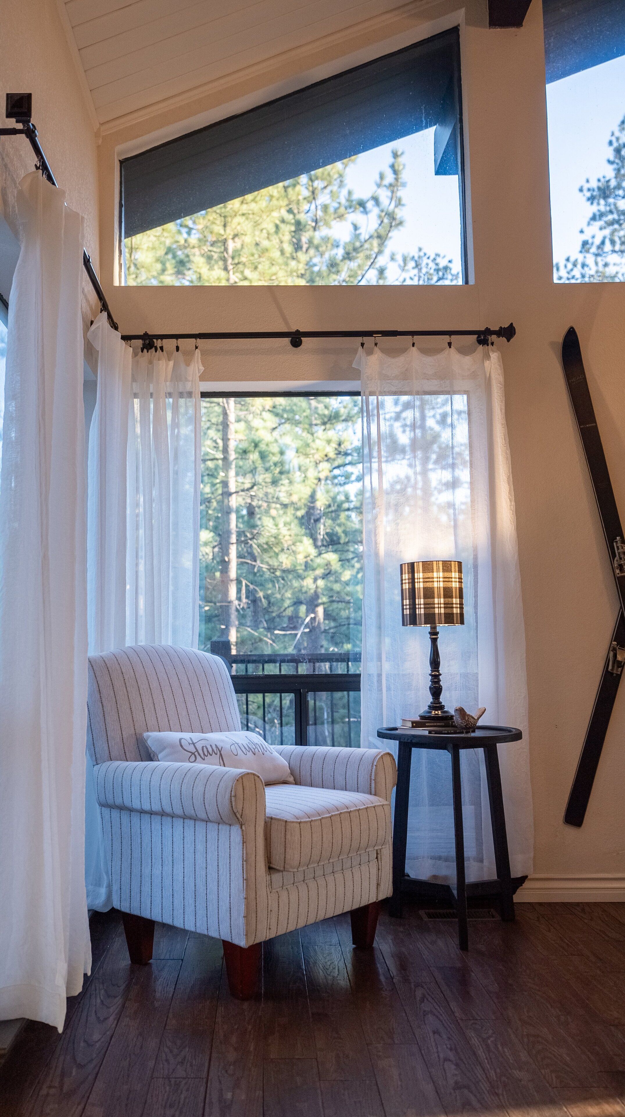 A cozy room with a large window, a chair, a small table with a lamp, and a ski board leaning against the wall.