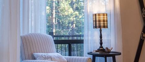 A cozy room with a large window, a chair, a small table with a lamp, and a ski board leaning against the wall.
